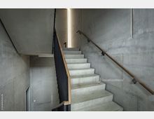 Betontreppe in einem modernen, minimalistisch gestalteten Innenraum mit Sichtbetonwänden und schwarzem Geländer. Hölzerne Handläufe und ein schmaler vertikaler Lichteinfall ergänzen das reduzierte Design.