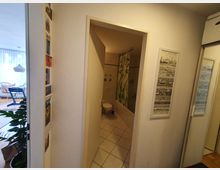 Flur mit Blick in ein Badezimmer. Das Badezimmer hat helle Fliesen, eine Badewanne mit Duschvorhang im Pflanzenmuster und eine Toilette. Links ist ein heller Wohnbereich mit großen Fenstern und einem weißen Lampenschirm sichtbar.