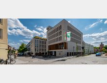 Moderne städtische Szenerie mit einem weißen, kubischen Gebäudekomplex, beschriftet als "Neues Rathaus Bernau", umgeben von Pflasterstraßen, Gehwegen und einer Fußgängerzone. Vor dem Gebäude stehen Flaggenmasten mit gehissten Fahnen, Fahrräder und einige Passanten; im Hintergrund sind weitere Gebäude und ein historischer Turm zu sehen.