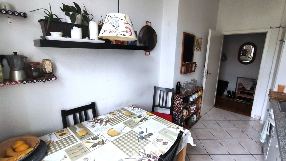 Small kitchen dining area with a rectangular table covered by a patterned tablecloth and black chairs, set against white walls with floating shelves holding plants, jars, and cookware. A tiled floor leads to an open doorway and a narrow sideboard with cups and glassware.