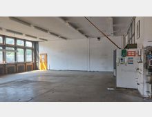 Leerer Industriebereich mit Betonboden, großen Fenstern auf der linken Seite, die viel Tageslicht hereinlassen, und einer orangefarbenen Tür. An der rechten Wand befinden sich Schaltkästen und Sicherheitseinrichtungen wie ein Verbandkasten und Spender.