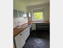 Kleine Küche mit L-förmiger Holzarbeitsplatte, weißer Spüle, Herd und weißen Unterschränken sowie Hängeschränken mit Glasfronten. Ein großes Fenster mit Rollladen bringt Tageslicht hinein; weiße Fliesenrückwand und dunkler, unregelmäßig gemusterter Steinboden.