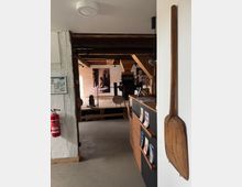 Innenansicht eines historischen Gebäudes mit Holzbalken, einem Infotisch mit Prospekten rechts und einem großen hölzernen Werkzeug an der Wand. Im Hintergrund sind Ausstellungsstücke und Schwarz-Weiß-Bilder sichtbar, vermutlich Teil eines Museums oder einer Ausstellung.