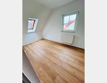 Leerer Dachgeschossraum mit weiß gestrichenen Wänden und schräger Decke, hellen Holzfußboden sowie zwei Fenstern (ein Dachfenster und ein senkrechtes Fenster). Unter dem rechten Fenster befindet sich ein Heizkörper.