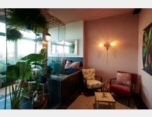 Gemütlicher Wohnraum mit rosa Wänden, dekorativer Beleuchtung und einem Mix aus modernen und klassischen Möbeln. Pflanzen, ein Glastrennwandbereich und ein kleines Holztischchen komplettieren die natürliche und einladende Atmosphäre.