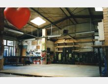 Ein Innenraum einer Werkstatt mit einer hohen Decke aus Metallträgern. Im Vordergrund hängt ein großes rotes Herz von der Decke, an den Wänden sind Regale, Werkzeuge und ein Wandbild einer Stadtsilhouette zu sehen.