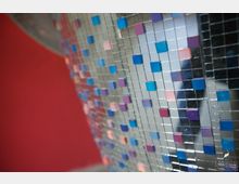 Nahaufnahme einer Fläche mit spiegelartigen Mosaikfliesen, die aus kleinen, glänzenden Quadraten in metallischem Silber sowie bunten Farben wie Blau, Rosa und Lila bestehen. Im Hintergrund ist eine rote Wand unscharf erkennbar.