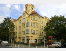 Gelbes, mehrstöckiges Wohn- oder Geschäftsgebäude im Stil der Gründerzeit mit markantem Erker und detailreichen Fensterelementen sowie einem kleinen Turm. Im Vordergrund befinden sich Bäume, ein Bürgersteig, parkende Fahrzeuge und eine Telefonzelle.