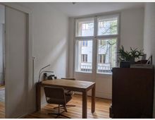 Ein kleiner Arbeitsraum mit einem Holzboden, einem schlichten Holztisch und zwei gepolsterten Stühlen. Große Fenster lassen Tageslicht herein und bieten Blick auf ein gegenüberliegendes Gebäude; auf einer Kommode stehen Zimmerpflanzen und Bücher.