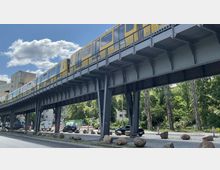 Gelbe Hochbahn der U-Bahn auf einer Stahlkonstruktion, die über eine Straße führt, mit Bäumen und parkenden Fahrzeugen im Hintergrund sowie einem teilweise bewölkten Himmel.