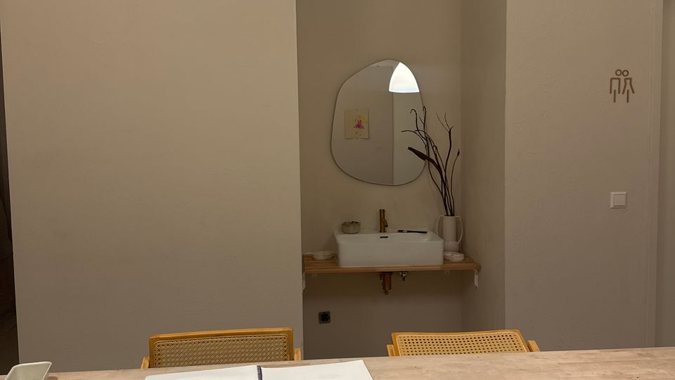 Moderner Innenraum mit langem Holztisch, darauf Zeichen- und Bastelmaterialien sowie Schalen; darüber hängt eine große weiße Pendelleuchte. Im Hintergrund ein kleines Waschbecken mit rundem Spiegel und ein Türbereich mit Toilettenpiktogramm an einer hellen Wand.