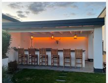 Überdachte Außenbar im Garten mit weißer Rückwand, dunkler Theke und sechs hohen Holz-Barhockern. Unter dem Dach hängen drei Laternenleuchten, und hinter der Theke sorgt eine orangefarbene indirekte Beleuchtung mit dekorativem Longhorn-Schädel an der Wand für Akzente.