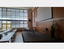 Großer Seminar- oder Konferenzraum mit hohen, rasterförmigen Fensterflächen und Jalousien sowie Reihen aus Tischen und Stühlen. Rechts befindet sich eine erhöhte Bühne mit Projektionsleinwand, Holzvertäfelung an der Wand und einzelnen Stühlen sowie einer Pflanzendekoration.