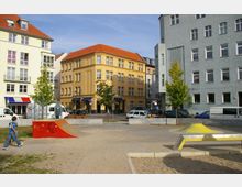 Eine kleine städtische Platzszene mit niedrigem Skatepark-Hindernis in Rot, umgeben von mehretagigen Wohnhäusern mit hellen Fassaden und roten Ziegeldächern. Im Hintergrund stehen geparkte Autos und Bäume, während der Platz von Sand- und Betonflächen geprägt ist.