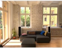 Wohnzimmer mit einer modernen Sitzlandschaft aus dunkelgrauem Stoff, hellem Holzboden und großen Fenstern mit Blick auf einen Innenhof. Die Wände bestehen aus weiß gestrichenen Ziegeln, und eine Topfpflanze steht in der Ecke neben der Balkontür.