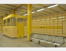 U-Bahn-Station mit gelb gefliesten Wänden, der Aufschrift "Weberwiese" und gelben Stützen. Im Vordergrund befinden sich eine Sitzreihe aus Metall und ein gelber Kabinenbereich mit Fenstern und Tür.