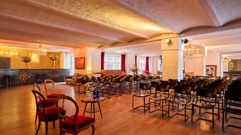 Großer Veranstaltungsraum mit Holzboden, niedriger Decke und weißen Stützpfeilern, bestuhlt mit mehreren Reihen schwarzer Stühle. Links befindet sich eine Bar mit dekorativer Wandgestaltung und Kronleuchtern, im Hintergrund große Fenster mit roten Vorhängen.