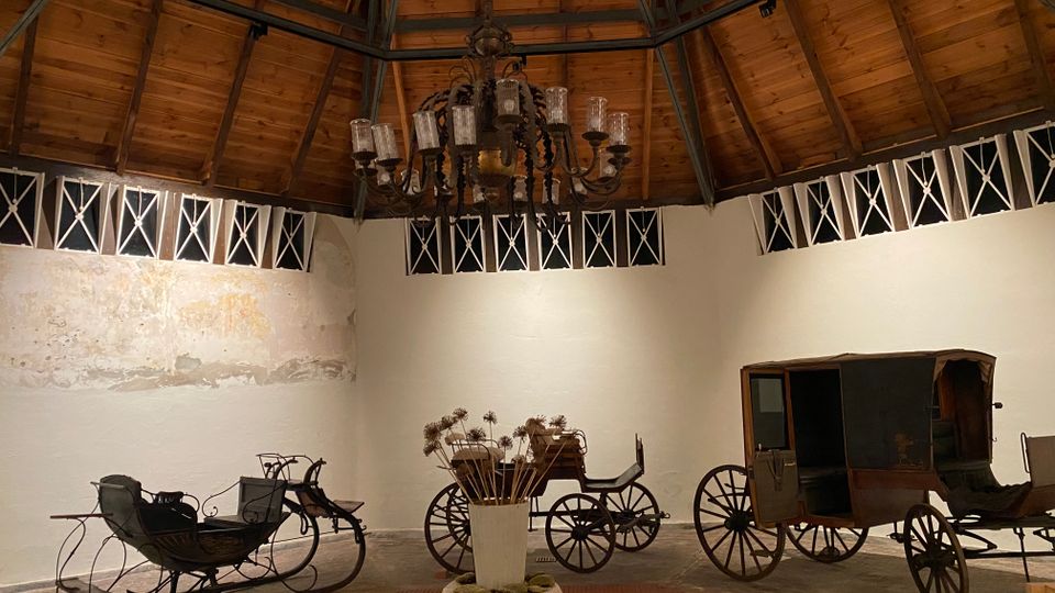 Innenraum einer runden Halle mit hohem Holzkuppeldach und sichtbaren Stahlträgern, in der Mitte ein großer Kronleuchter. An den weißen Wänden mit umlaufenden Fensteröffnungen stehen mehrere historische Pferdekutschen und Schlitten ausgestellt.