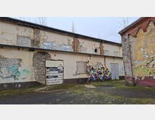 Verwitterter Hinterhof mit einer langen, flachen Gebäudefassade aus bröckelndem Putz und freiliegendem Mauerwerk, teils zugemauerten Öffnungen und Graffiti. Rechts steht ein weiteres Gebäude mit abplatzender Farbe; der Boden wirkt rissig und moosbewachsen.