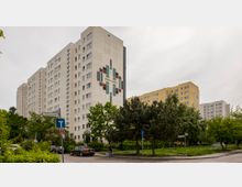 Mehrstöckige Wohnblöcke mit schlichten Fassaden, von denen einer an der Giebelwand ein großes, geometrisches Wandmosaik zeigt. Im Vordergrund befinden sich eine Kreuzung, ein Straßenschild, geparkte Autos sowie dichte Bäume und Grünflächen.