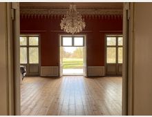 Großer historischer Saal mit Holzdielenboden, dunkelroten Wänden und Stuckfries an der Decke, in der Mitte ein Kristallkronleuchter. Drei hohe Fenstertüren, die mittlere geöffnet, führen auf eine Grünfläche im Außenbereich.