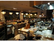 Eine offene, moderne Restaurantküche mit Edelstahl-Arbeitsflächen, Kochausrüstung und zahlreichen Zutaten in Plastikbehältern. Über der Küche hängen runde Deckenlampen, und im Hintergrund sind Fenster und Getränkeflaschen sichtbar.