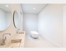 Modernes Badezimmer mit minimalistischem Design, bestehend aus einer freistehenden weißen Badewanne, einem Waschtisch mit weiß-grauer Marmorplatte und zwei eingelassenen Waschbecken. An der Wand befinden sich zwei runde Spiegel, und ein kleines Fenster lässt Tageslicht herein.