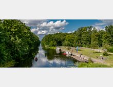 Ein Wasserkanal, umgeben von dichten Bäumen, mit einem kleinen Anlegesteg auf der rechten Seite, an dem Boote festgemacht sind und Menschen sich aufhalten. Im Vordergrund fährt ein Ruderboot, und im Hintergrund erstreckt sich der Kanal in die Ferne unter einem bewölkten, blauen Himmel.