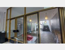 Eingangsbereich eines Gebäudes mit einer Glastür in einem goldenen Rahmen. Im Inneren befinden sich Hängelampen, eine Treppe mit Metallgeländer, gestapelte Stühle an der rechten Wand sowie ein roter Teppich und Glasflächen, die eine helle Atmosphäre schaffen.