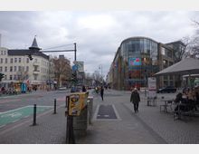 Straßenszene in einer urbanen Umgebung mit einer Kreuzung; links ein älteres Gebäude mit spitzem Turm und Erdgeschossgeschäften, rechts ein modernes Gebäude mit runder Glasfassade und einem "Sale"-Schild. Im Vordergrund Gehwege, ein Fahrradstreifen, Fußgänger, ein Straßenschild mit S-Bahn-Symbol und ein Café mit Sitzgelegenheiten.