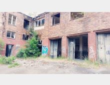 Backsteinfassade eines verlassenen Industriegebäudes mit mehreren eingeschlagenen Fenstern und offenen Holztoren. Die Umgebung ist überwuchert von Gras und Sträuchern, mit Graffiti an den Wänden.