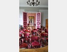 Ein historischer Innenraum mit einer Anordnung von roten Holzstühlen auf einem gemusterten Teppich. Im Hintergrund sind geöffnete, rot-weiße Doppeltüren zu sehen, die den Blick auf mehrere hintereinanderliegende Räume mit Kronleuchtern freigeben.