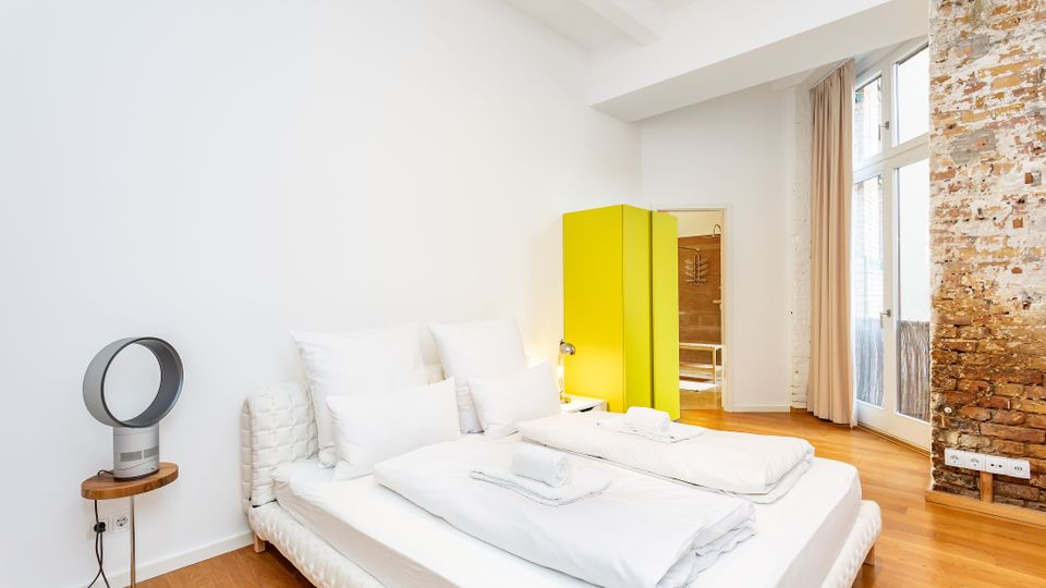 Helles, minimalistisches Schlafzimmer mit Doppelbett und weißer Bettwäsche auf Holzboden, neben einem kleinen Beistelltisch mit Ventilator. Im Hintergrund stehen ein auffällig gelber Kleiderschrank und hohe Fenster mit Vorhängen neben einer freigelegten Ziegelwand.