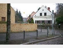 Straßenszene mit einem weißen Fachwerkhaus mit dunklen Fensterläden und einem Ziegeldach im Hintergrund. Im Vordergrund sind eine gepflasterte Straße, eine niedrige gelbe Backsteinmauer und einzelne Bäume zu sehen.