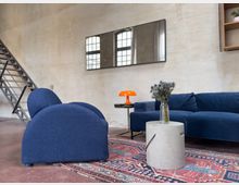 Wohnzimmer mit industriellem Design, ausgestattet mit einem blauen Sessel und Sofa, einem gemusterten Teppich, einem Beton-Beistelltisch mit Vase, und einer orangefarbenen Lampe. Im Hintergrund eine Metalltreppe und große Fenster mit schwarzem Rahmen.