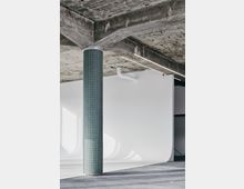 Industrieähnlicher Innenraum mit einer freistehenden Säule, die mit kleinen grünen Fliesen verkleidet ist, und einer groben Betondecke. Weiße, abgerundete Wände und ein glatter Boden erzeugen einen minimalistischen Kontrast.