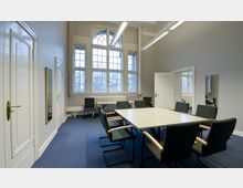 Besprechungsraum mit einem rechteckigen Tisch und mehreren gepolsterten Stühlen, großen Fenstern mit Sprossen, einem Heizkörper unter dem Fenster und hellgrauen Wänden. Die Decke ist mit langen, rechteckigen Deckenleuchten ausgestattet, und eine Tür sowie ein Spiegel befinden sich an der linken Wand.