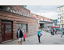 Straßenszene in einer Stadt mit einem Backsteingebäude im Vordergrund, einer Hochbahnbrücke mit fahrender S-Bahn darüber und einem U-Bahn-Zugang der Linie U5 rechts im Bild. Mehrere Menschen gehen über den Gehweg, während Fahrräder und Autos im Hintergrund zu sehen sind.