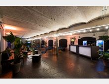 Industriell gestalteter Veranstaltungsraum mit abgerundeter Backsteindecke, minimalistischen Möbeln und großen Pflanzen. Der Raum verfügt über eine beleuchtete Bar, Kühlschränke und Sitzbereiche mit blauen Stühlen und Tischen.