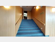 Treppe mit blauem Teppichboden, flankiert von Holzwänden und Handläufen, führt zu einer geschlossenen Tür. Wandleuchten und eine Beschilderung mit der Aufschrift "13–19" sind sichtbar.