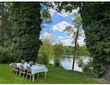 Ein Holztisch mit sechs Stühlen steht auf einer grünen Wiese zwischen zwei hohen, dicht bewachsenen Bäumen. Im Hintergrund ist ein See mit umliegenden Bäumen und blauem Himmel zu sehen, sowie eine Lichterkette, die zwischen den Bäumen gespannt ist.