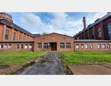 Backstein-Gebäude mit flachem Dach und mehreren Fenstern, flankiert von großen, mehrstöckigen Industriegebäuden ebenfalls aus rotem Backstein. Im Vordergrund ein gepflasterter Weg und eine Wiese, im Hintergrund ein Schornstein und bewölkter Himmel.