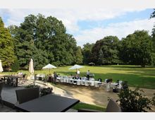 Große Gartenanlage mit einer freien Rasenfläche, umgeben von hohen Bäumen. Im Vordergrund stehen gedeckte Tische und Stühle unter Sonnenschirmen, während Menschen die Umgebung auf einer Terrasse oder im Garten genießen.