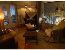 Gemütliches Wohnzimmer mit Holzboden, gedämpfter Beleuchtung und einer Einrichtung im Vintage-Stil. Im Vordergrund stehen Sessel, ein Hocker und ein Schaukelstuhl, mit einem großen Fenster im Hintergrund, geschmückt mit hängenden Pflanzen und Dekorationslampen.