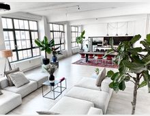 Eine moderne, helle Loftwohnung mit großen Industrie-Fenstern, einem offenen Wohn- und Essbereich. Im Vordergrund befinden sich beige Sofas und Pflanzen, im Hintergrund ein langer Esstisch mit roten Stühlen auf einem Teppich sowie eine offene Küche mit schwarzen Fliesen.