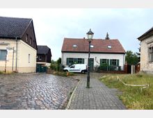 Gepflasterte Dorfstraße mit mehreren Wohnhäusern, darunter ein weiß verputztes Haus mit grünen Fensterläden und rotem Ziegeldach. Im Vordergrund eine Straßenlaterne und ein geparkter weißer Kleintransporter, daneben eine kleine Grünfläche.