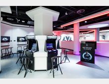 Eine moderne Gaming-Lounge mit Arcade-Spielautomaten, stilisierten schwarzen Hockern und Bildschirmen an den Wänden. Der Raum ist hell beleuchtet mit weißen und pinkfarbenen LED-Lichtern, und ein großes Logo mit der Aufschrift „AVA“ ist im Hintergrund sichtbar.