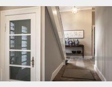 Flur eines Hauses mit einer weiß lackierten Treppe und einer Glastür auf der linken Seite. Im Hintergrund steht ein Tisch mit blau-weißen Porzellanvasen und einer gerahmten Wanddekoration darüber; hölzerner Fischgrätboden und eine sternförmige Hängelampe ergänzen den Raum.