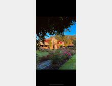 Ein ländliches Gebäude mit Backsteinfassade, umgeben von einem Garten mit Blumenbeeten voller Rosen und gepflegtem Rasen. Im Hintergrund stehen Bäume, und die Szene wird teilweise von hängenden Zweigen eingerahmt.