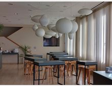 Ein moderner, heller Innenraum mit hohen Fenstern und transparenten Vorhängen, ausgestattet mit hohen Tischen und Holzbarhockern. Große, weiße, kugelförmige Hängelampen dominieren die Decke, während eine Treppe an der linken Wand und ein schwarzer Bildschirm im Hintergrund sichtbar sind.